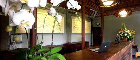 A wooden reception desk with a laptop, potted orchids, and artwork on the wall.