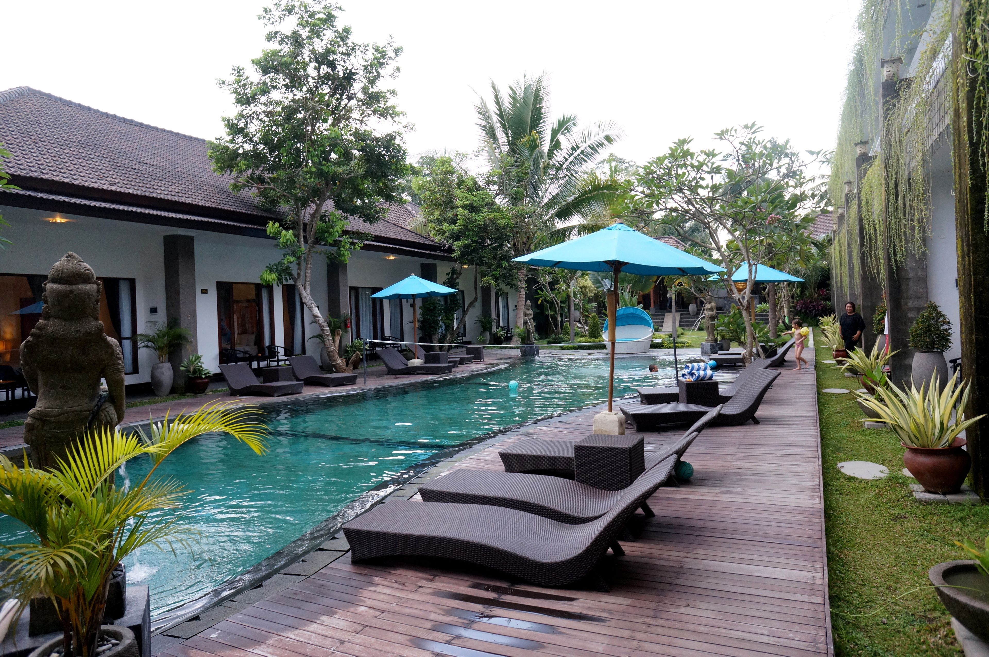 A pool area with lounge chairs and umbrellas, surrounded by greenery and a building with a tiled roof.