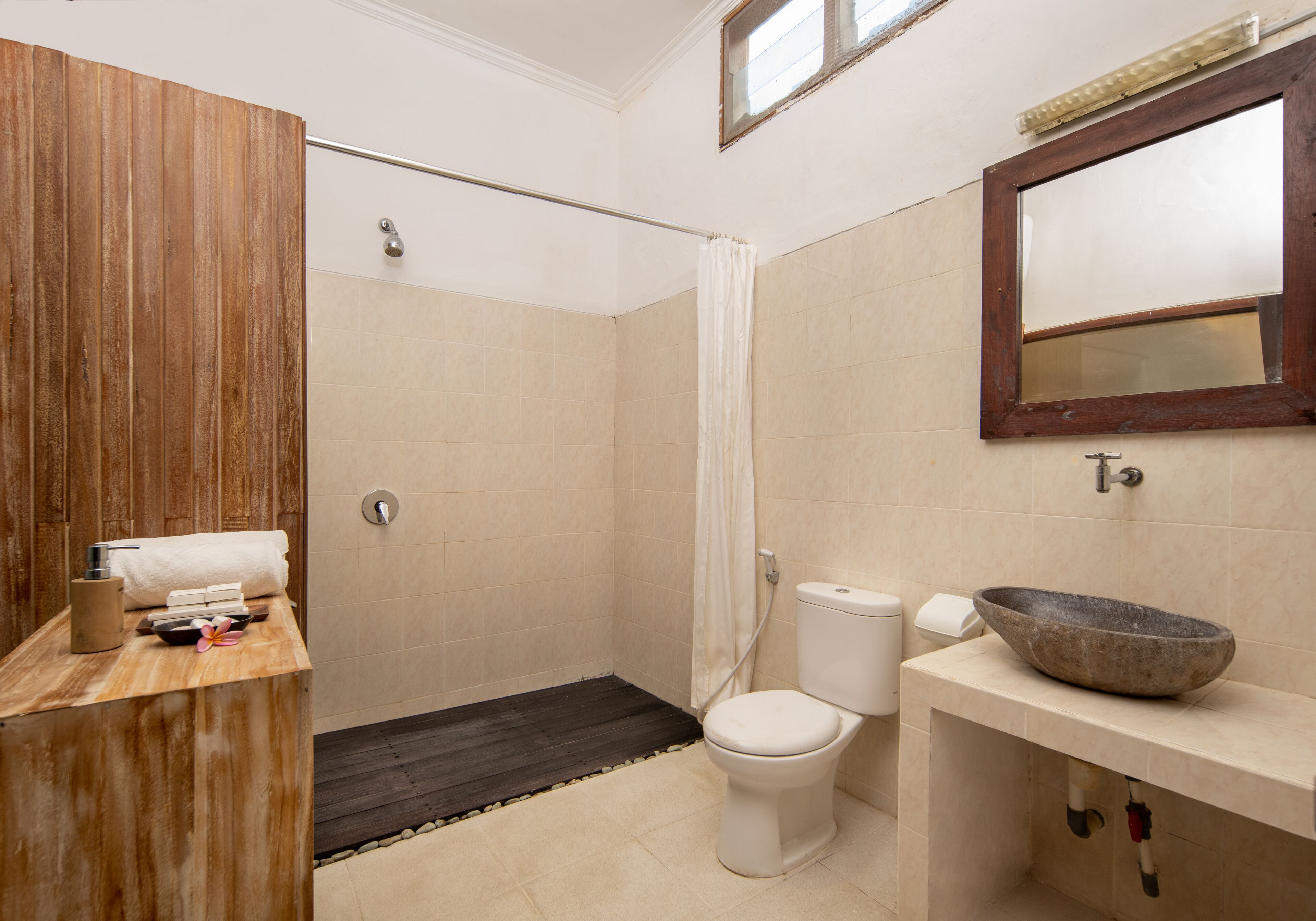 Une salle de bain avec un meuble-lavabo en bois, des toilettes blanches, un lavabo en pierre et une douche dotée d’une porte en bois.