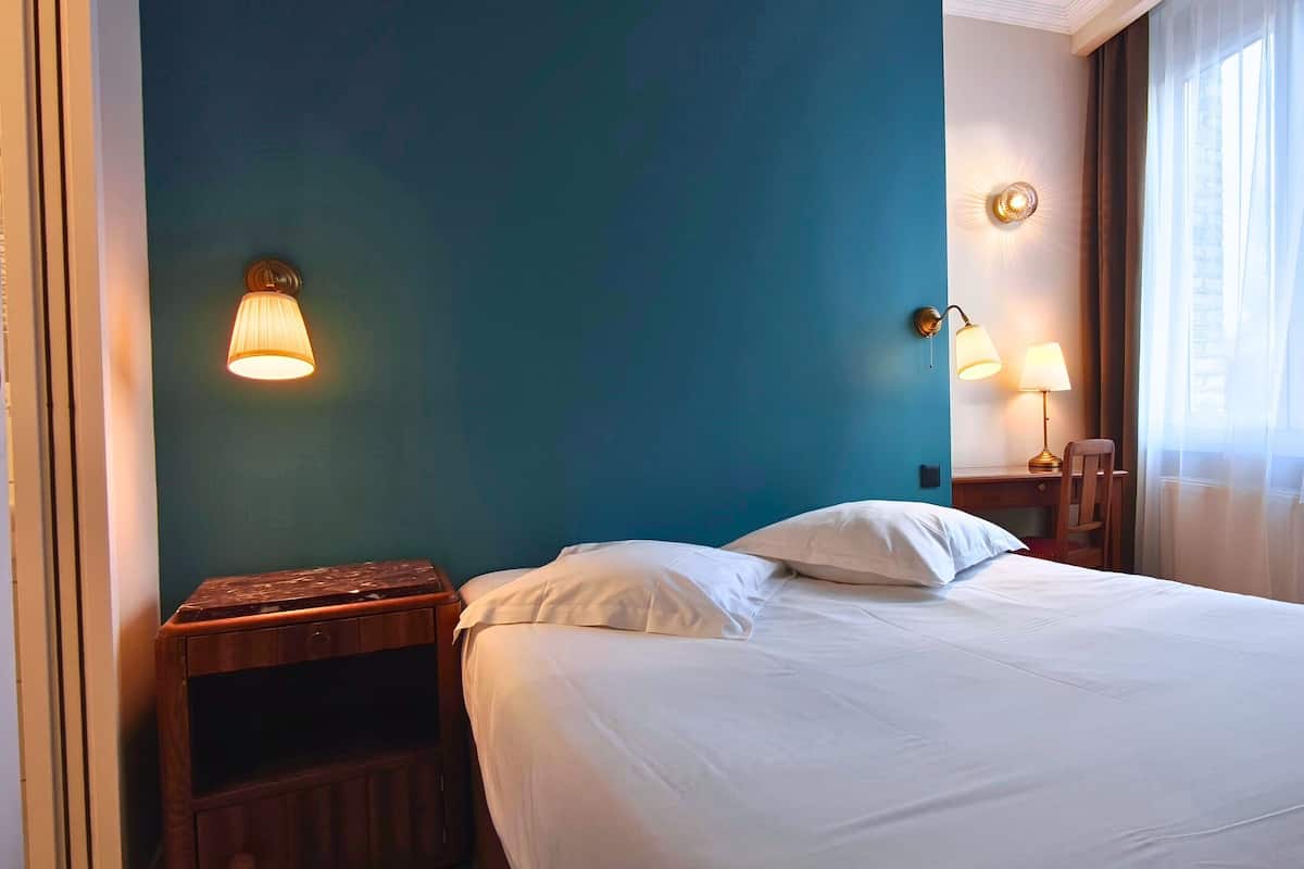A hotel room with a bed, two bedside lamps, a nightstand, and a window with curtains.