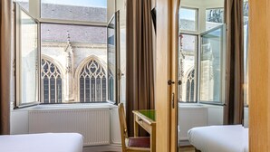 A room with two beds, a wooden chair, and a view of a building with arched windows. - Hotel Saint Maurice (Lille)