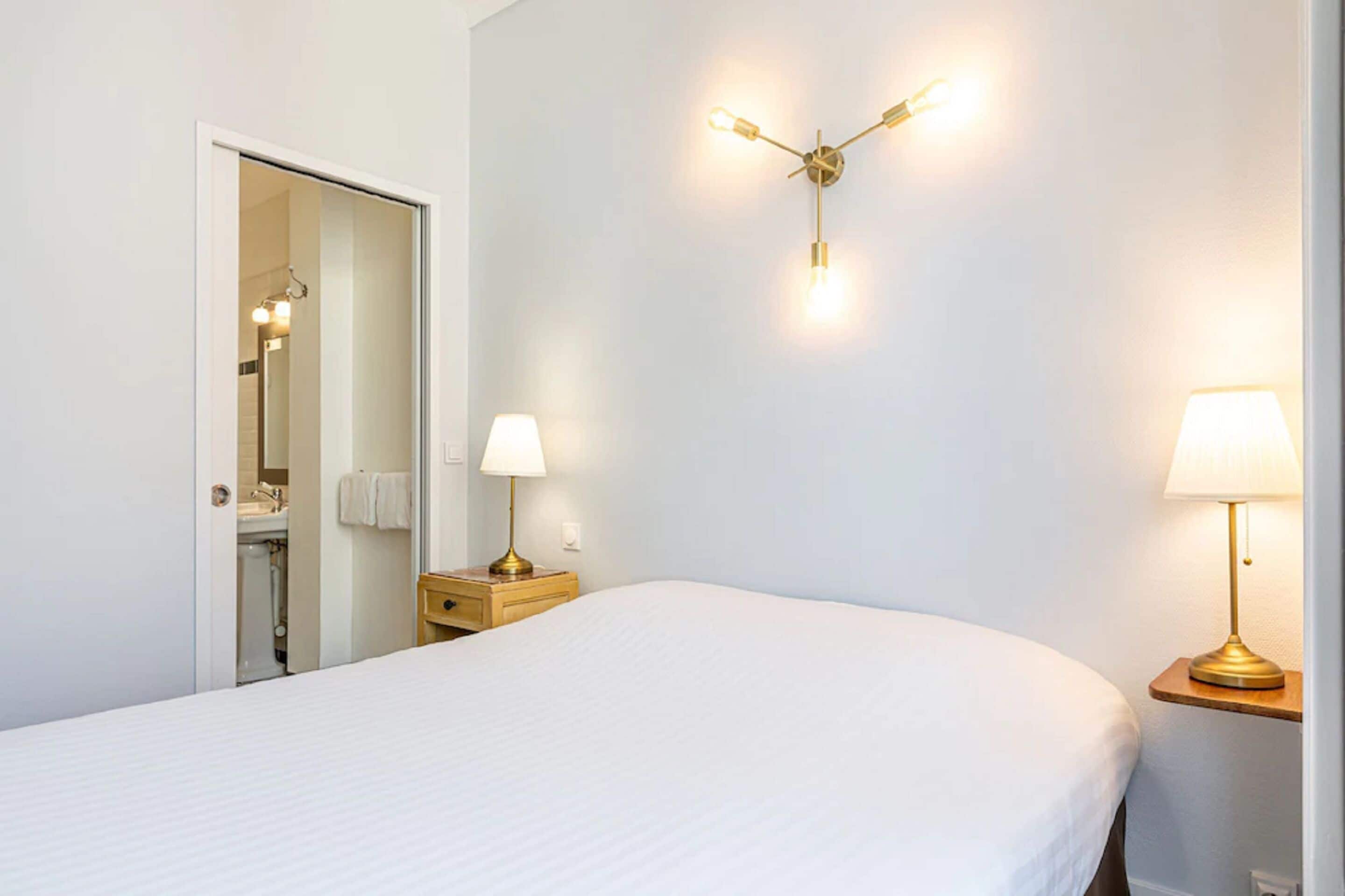A hotel room with a bed, two bedside tables with lamps, a wall-mounted light fixture, and a door leading to a bathroom.