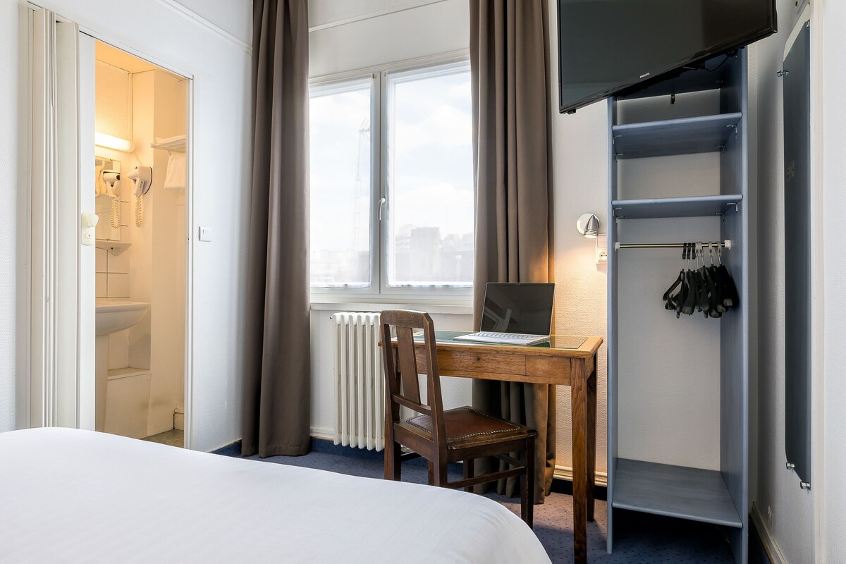 Una habitación de hotel con una cama, un escritorio de madera con una computadora portátil, una silla, un radiador, una ventana con cortinas y un armario con ropa colgada.