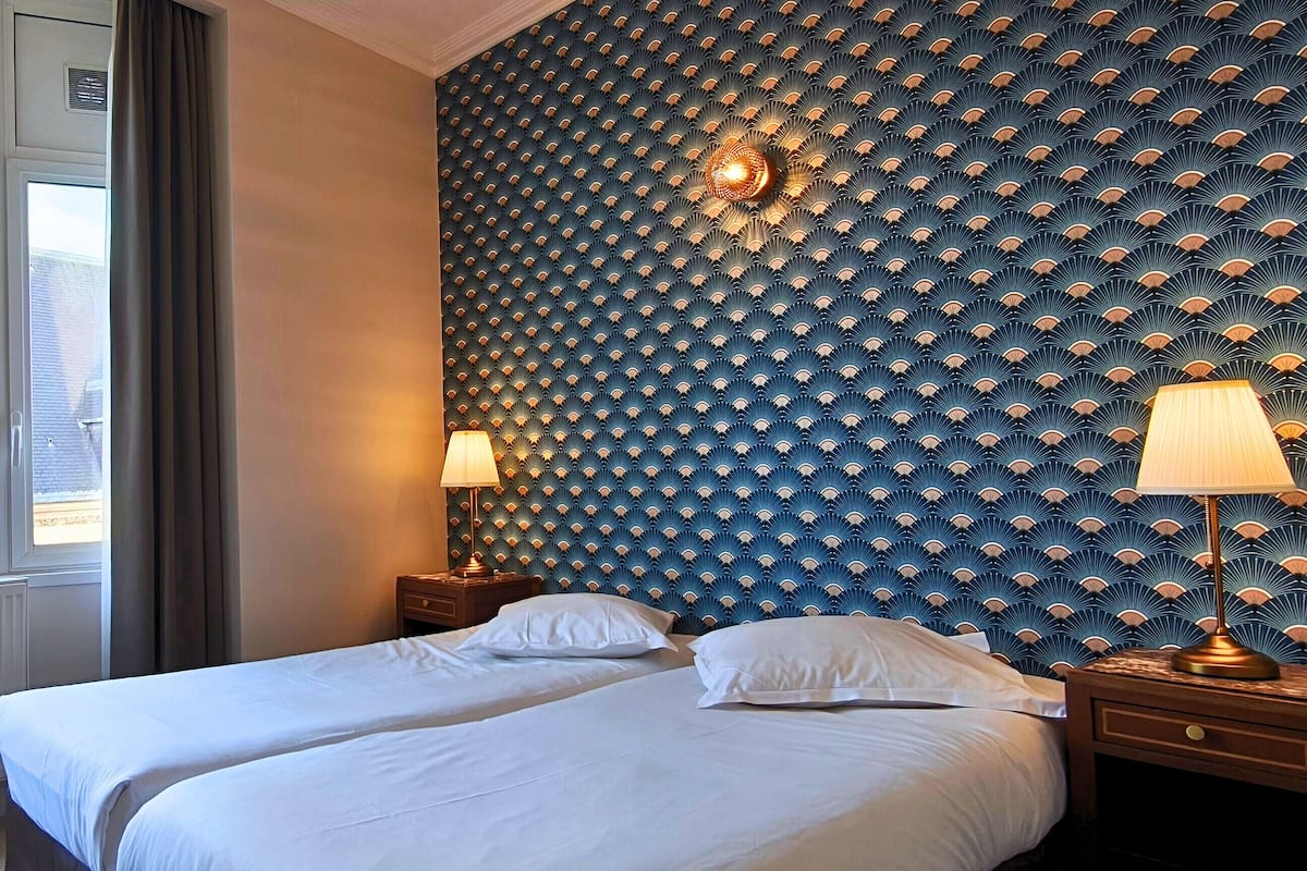 A hotel room with two beds, a large window with a view of a building, and a patterned wall.
