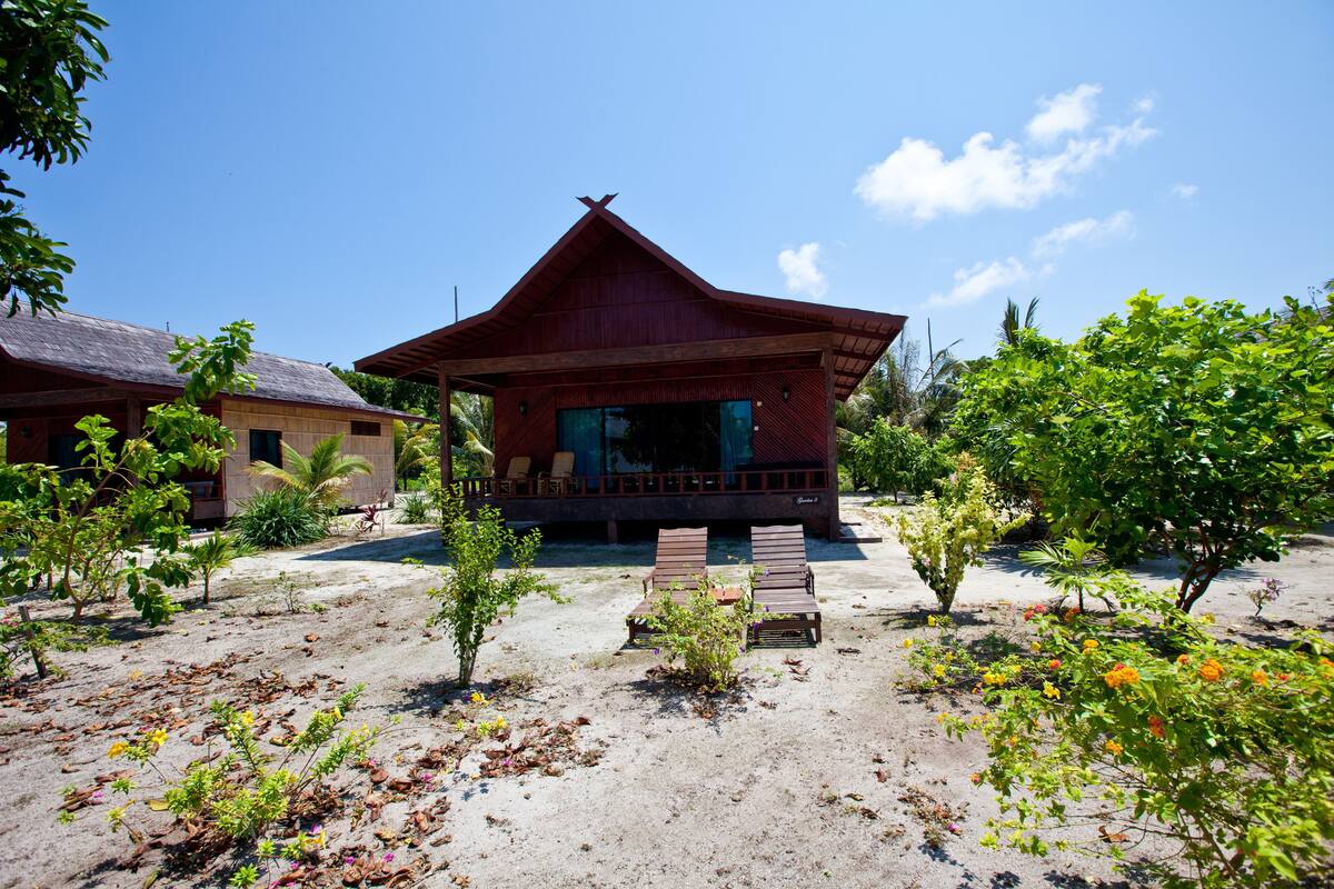 A modern house with a large porch, surrounded by greenery and a sandy ground.