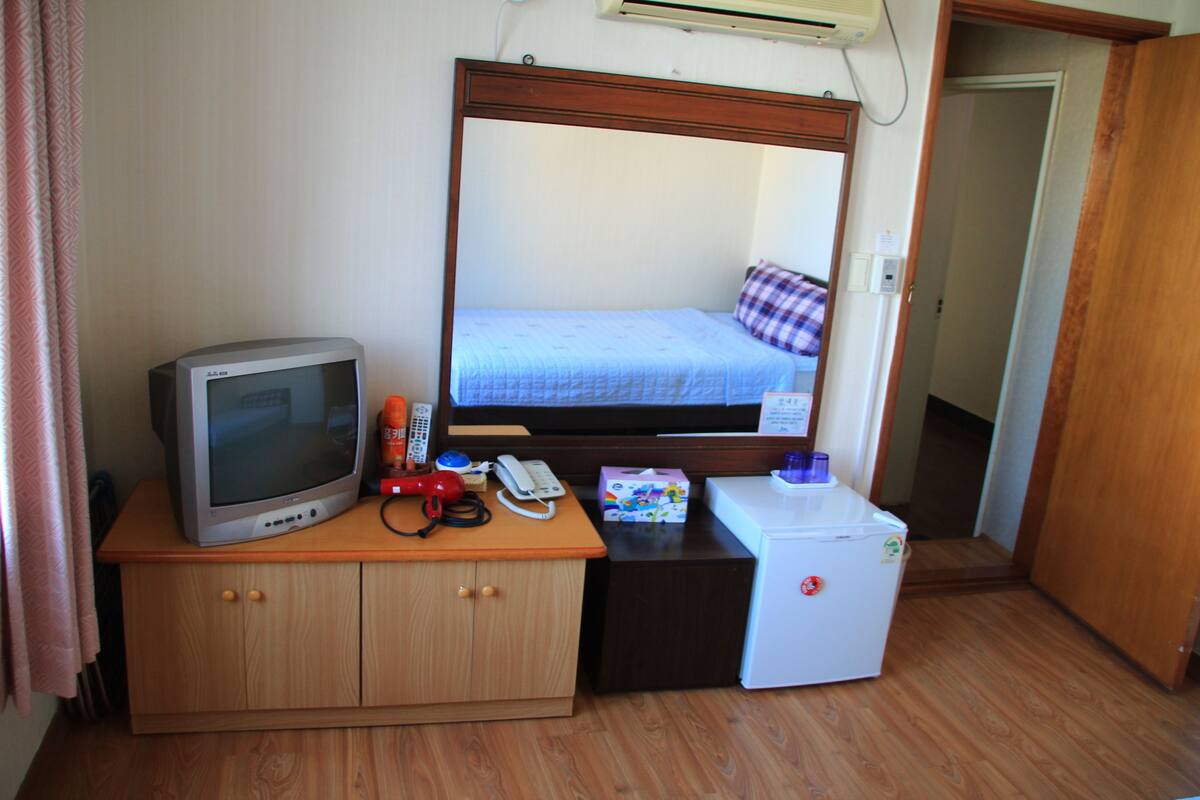 A compact room with a bed, a small desk with a television, a refrigerator, and a telephone.