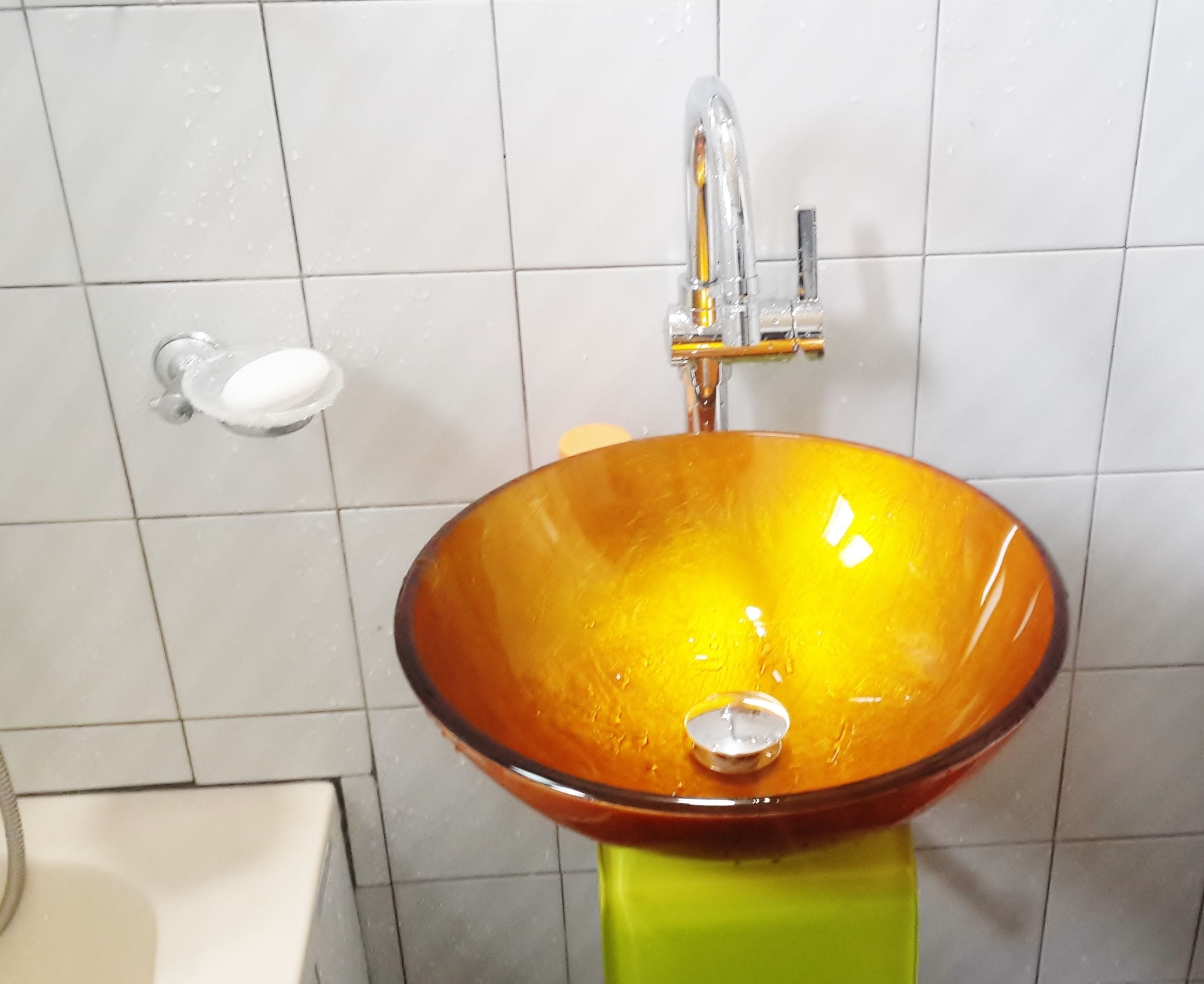 A bathroom with a glass bowl sink, a green pedestal, and a white tiled wall.