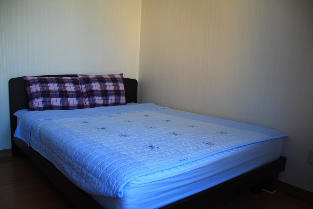 A single bed with a blue quilt and two plaid pillows, a wooden headboard, and a plain wall.
