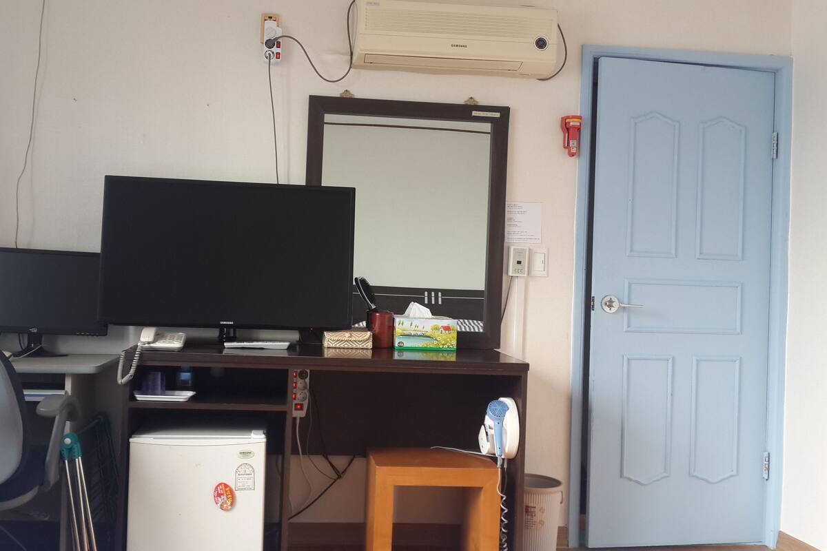 A small office with two monitors, a desk, a chair, a refrigerator, a blue door, and an air conditioner.