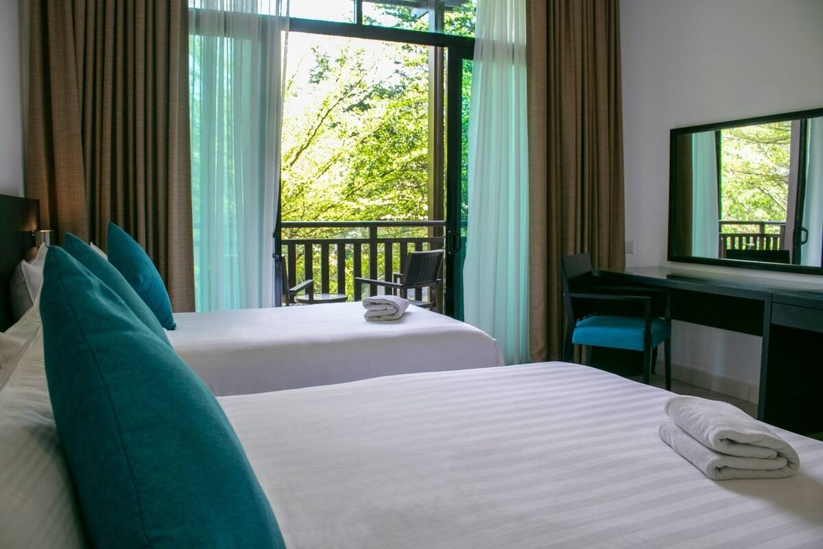 A hotel room with two beds, a desk, and a balcony with a view of greenery.
