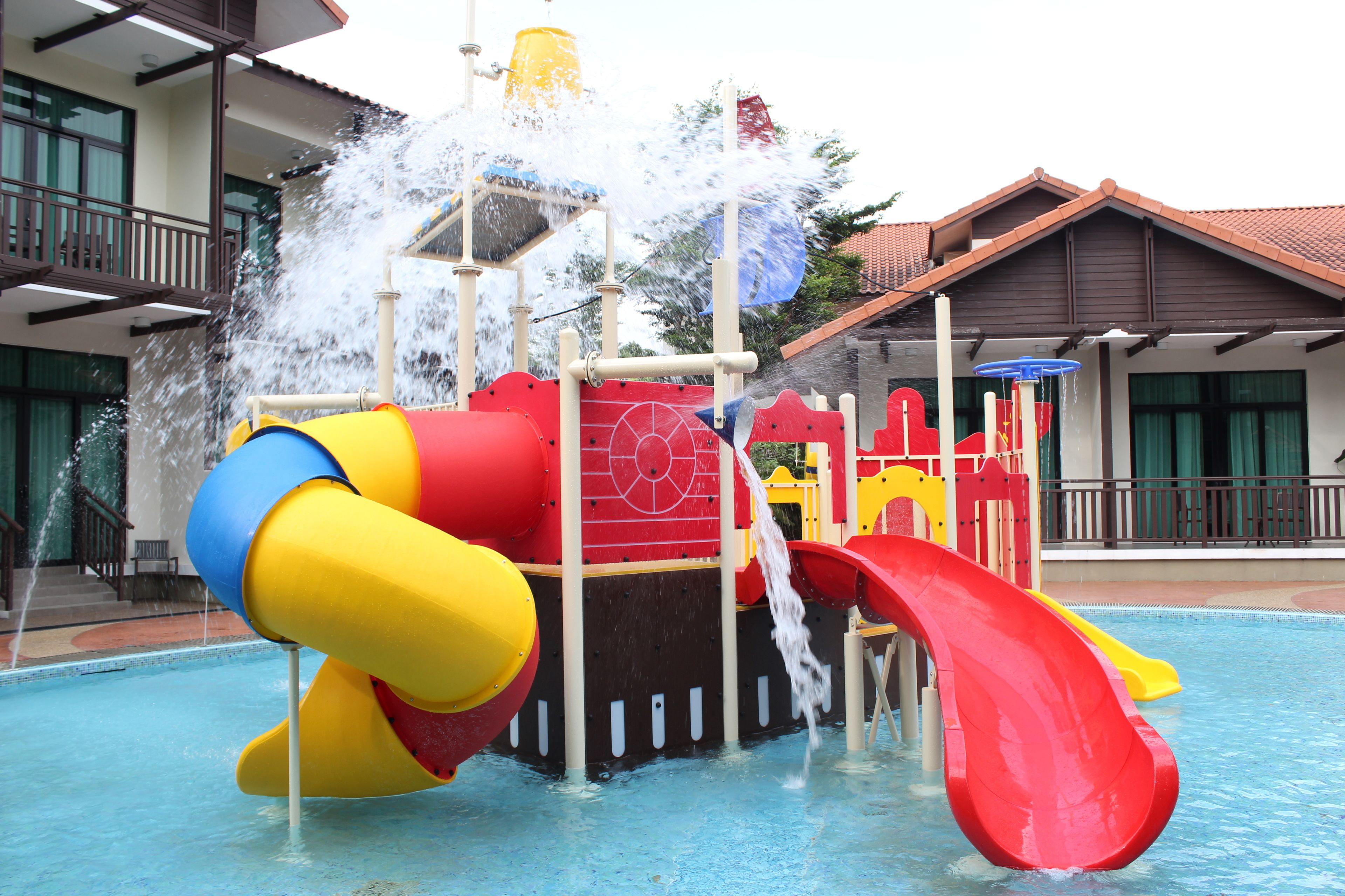 A water play area with slides and water jets, adjacent to a swimming pool, with buildings in the background.