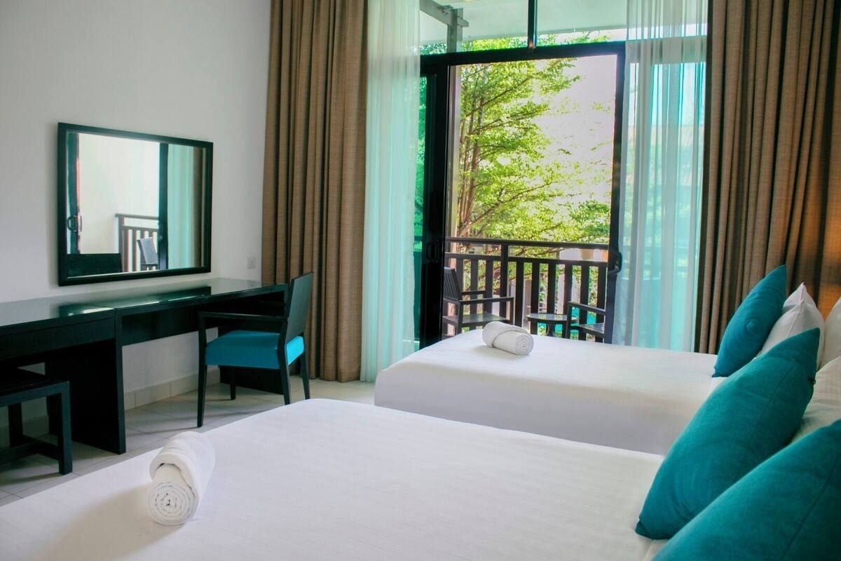 A hotel room with two beds, a desk, a chair, and a large window with a view of greenery.