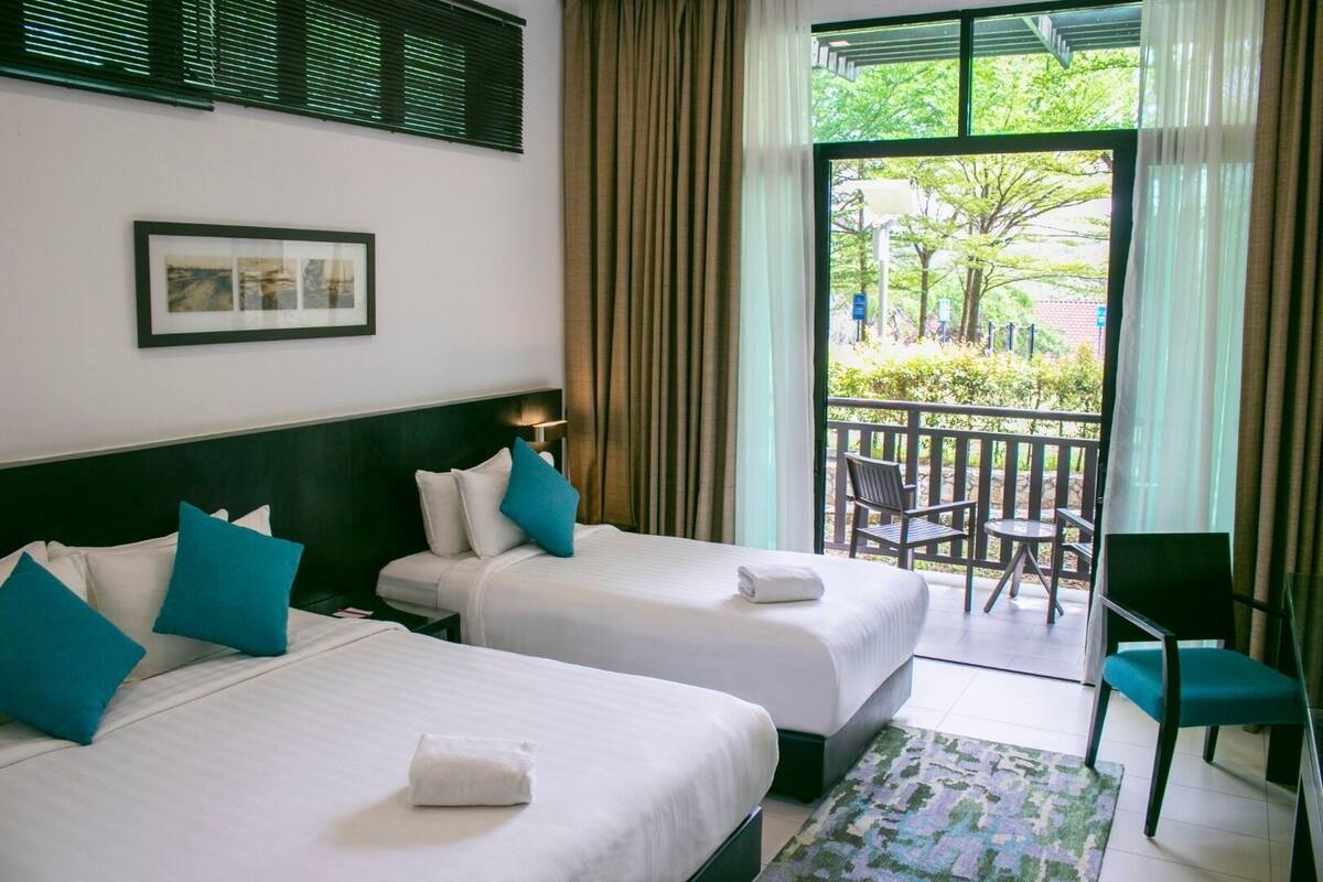 A hotel room with two beds, a balcony with chairs, and a view of greenery.