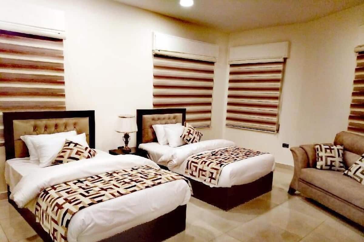 A hotel room with two beds, a sofa, and large windows with blinds.