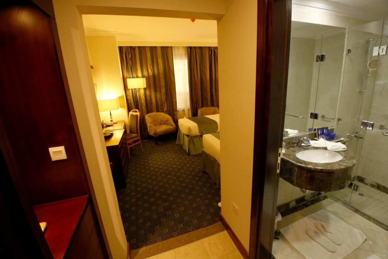 Habitación de hotel con cama, una silla, un escritorio con lámpara, y baño con lavabo y ducha.