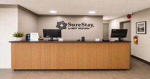 Uma mesa de recepção com uma placa indicando 'SureStay by BEST WESTERN', dois monitores, uma planta e um extintor de incêndio.