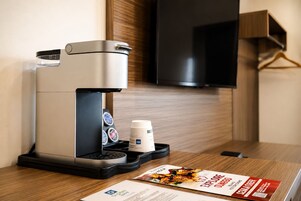 Una cafetera con espumador de leche y un vaso desechable sobre una mesa de madera en una habitación de hotel.