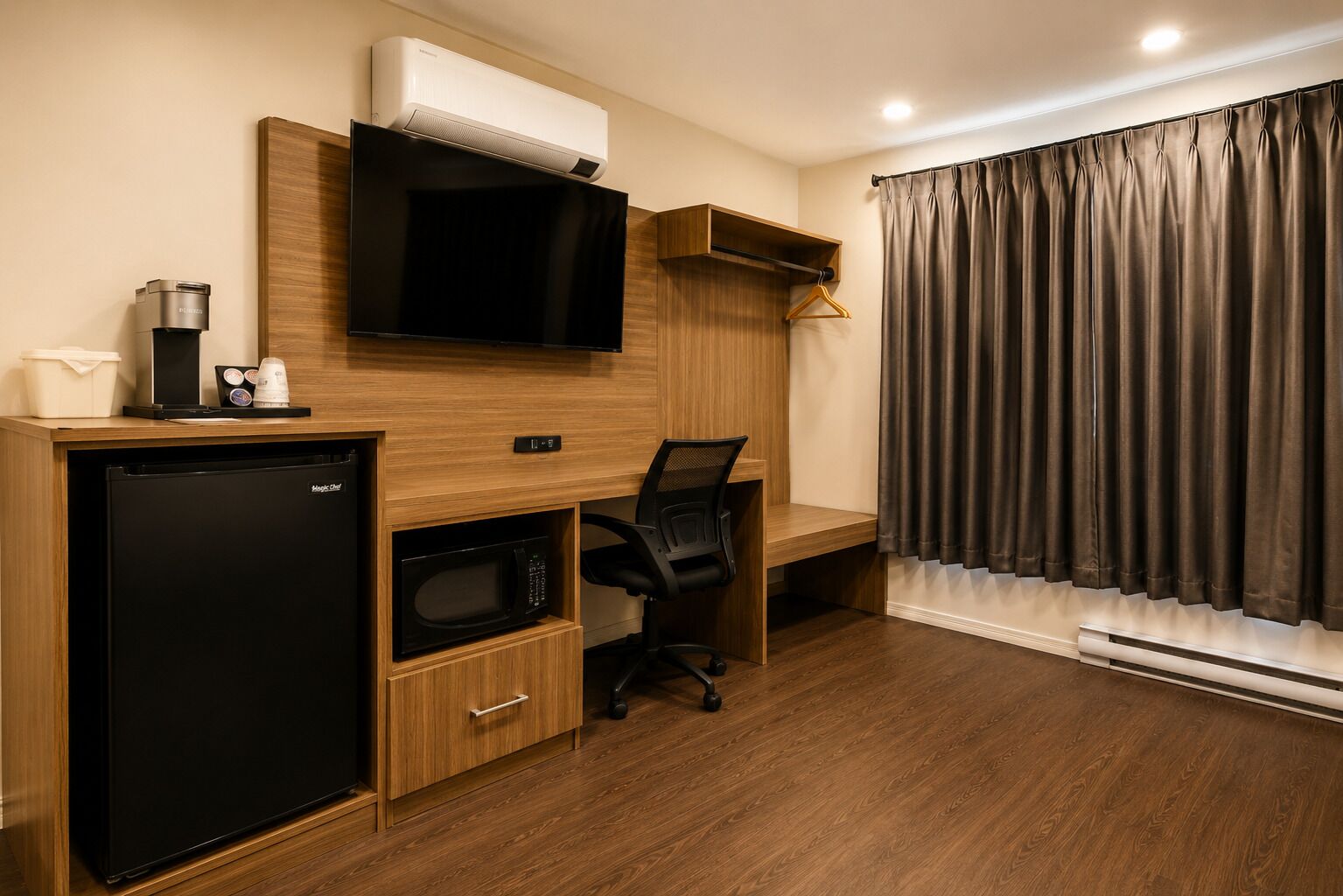 Une chambre d’hôtel compacte dotée d’une kitchenette, d’un téléviseur à écran plat, d’un bureau en bois, d’une chaise et d’une grande fenêtre avec des rideaux.