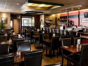 Interior de un restaurante con varias mesas y sillas, una zona de bar con mostrador y estantes llenos de botellas, y un ventanal que proporciona luz natural.