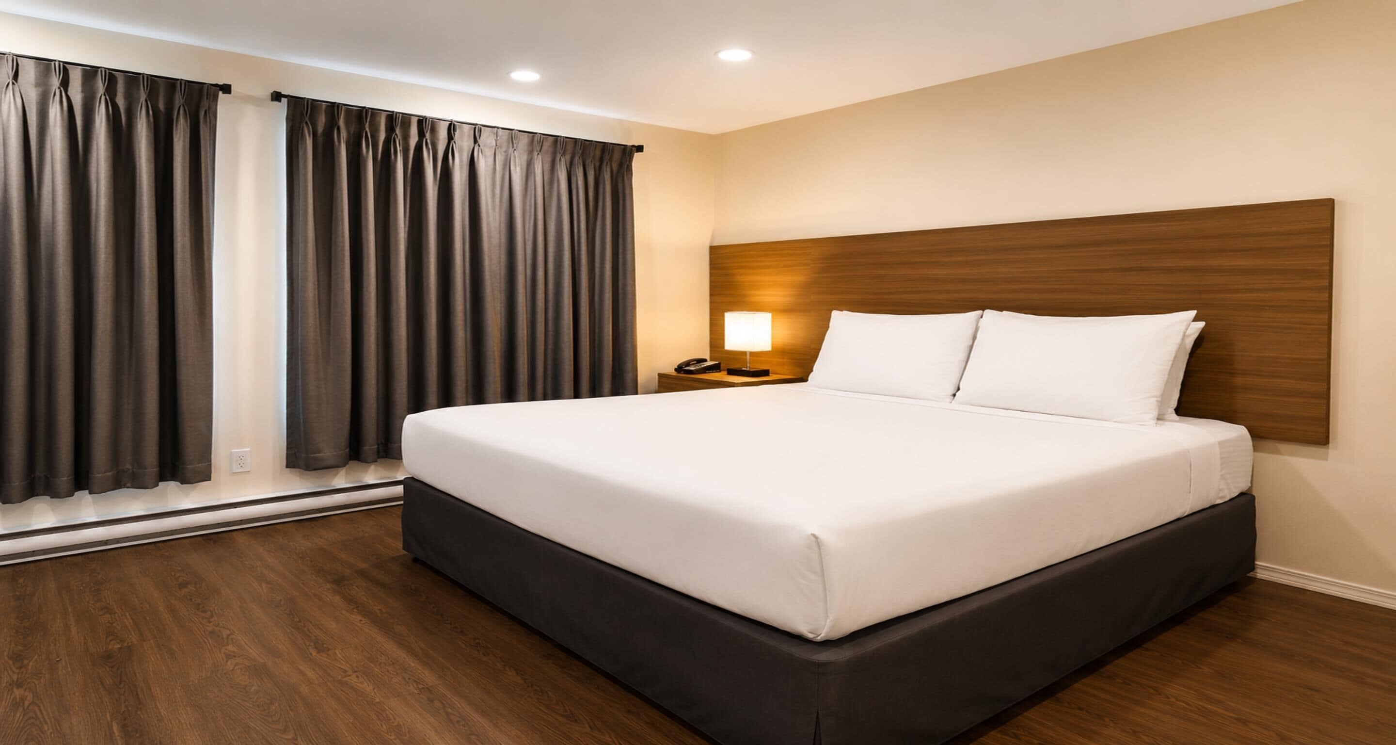 A hotel room with a large bed, wooden headboard, a bedside table with a lamp, and a window with curtains.
