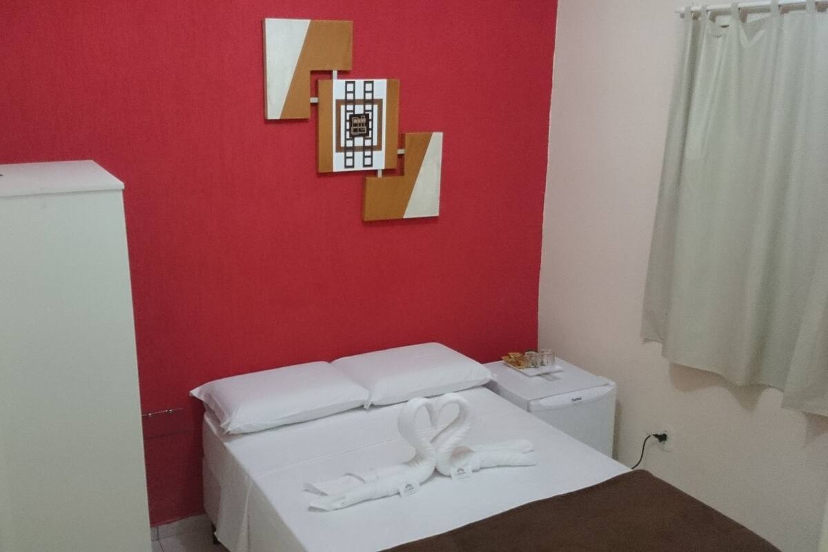 A small, simple room with a bed, a nightstand, and a red wall with a geometric decoration.