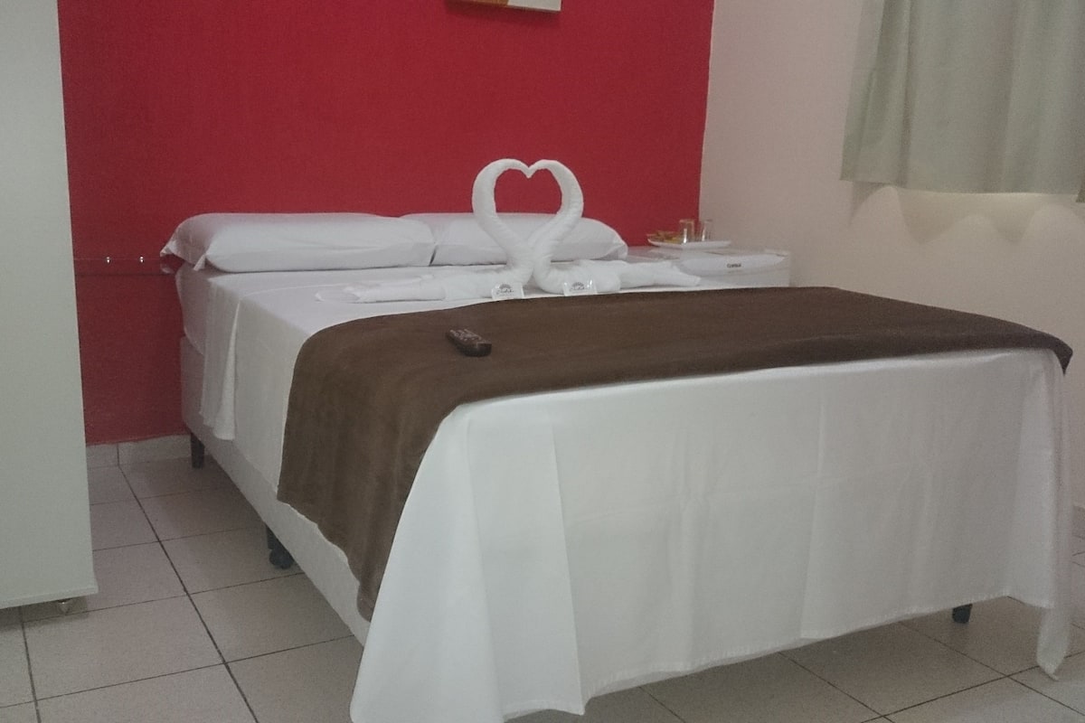 A hotel room with a bed, a brown blanket, a heart-shaped towel, and a remote control on the bed.