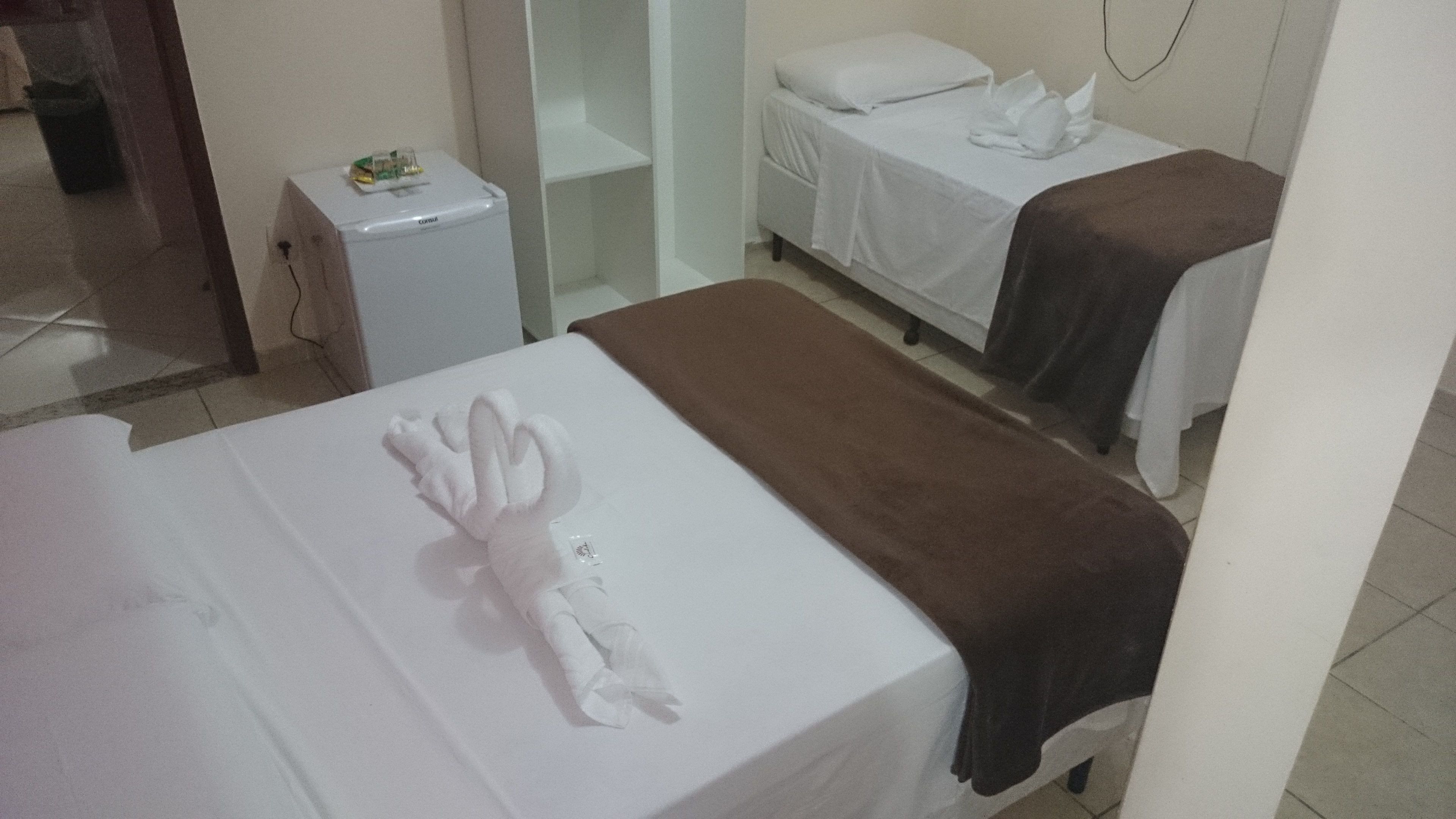 A hotel room with a bed, a small white refrigerator, a bedside table with a towel, and a brown blanket on another bed.