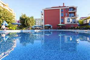Ein Schwimmbecken mit Fliesenboden, umgeben von Sonnenschirmen und Liegestühlen, mit Gebäuden im Hintergrund.