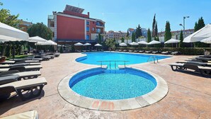 Una piscina con sillas de descanso y sombrillas, rodeada por un edificio de varios pisos y fachada roja.