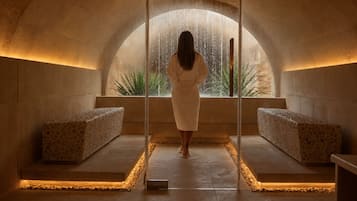 Un espacio de spa con una mujer de pie frente a un gran ventanal arqueado, dos bancos y un suelo de piedra.