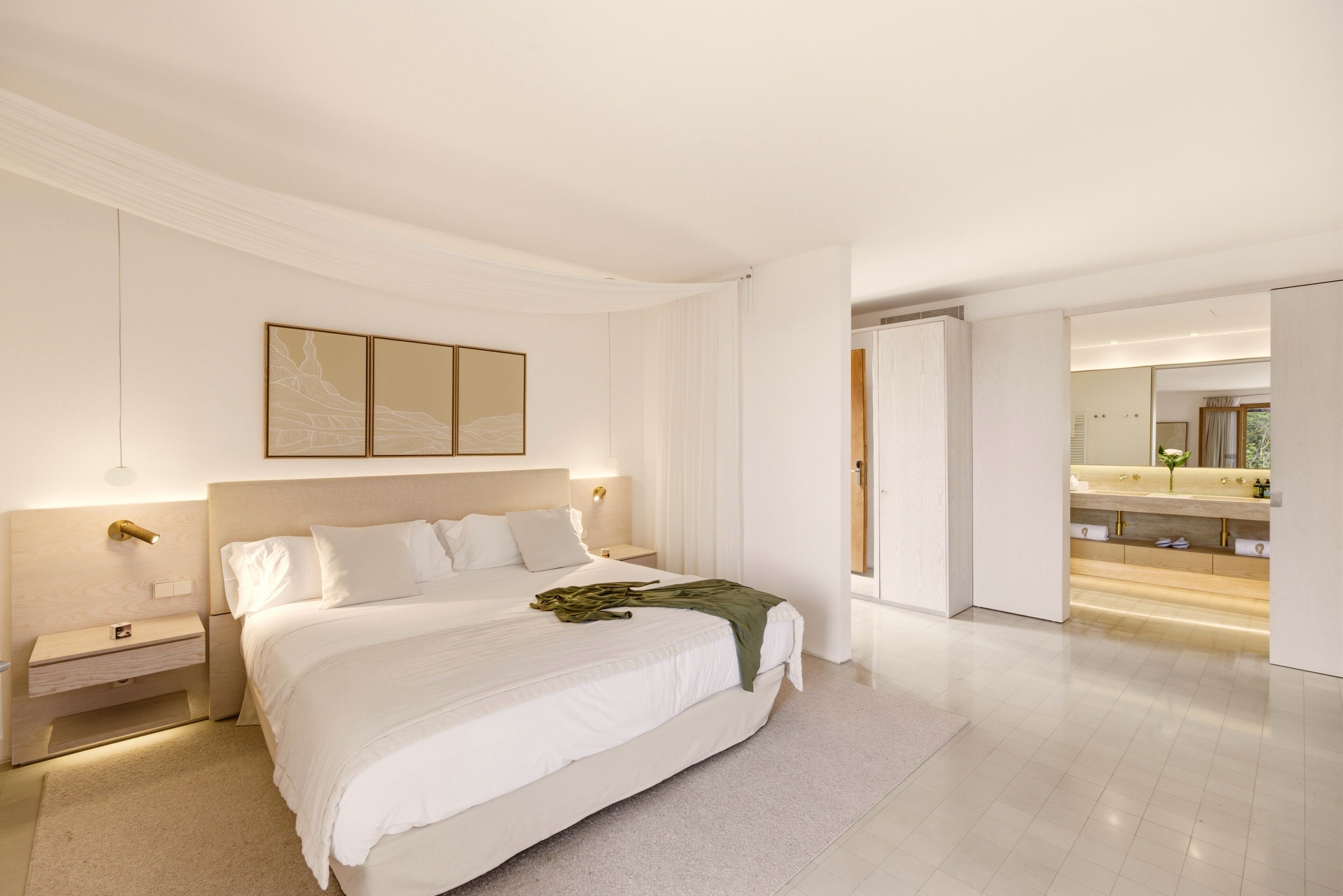 A modern bedroom with a large bed, bedside tables, a nightstand, a mirror, and a bathroom with a vanity and sink.