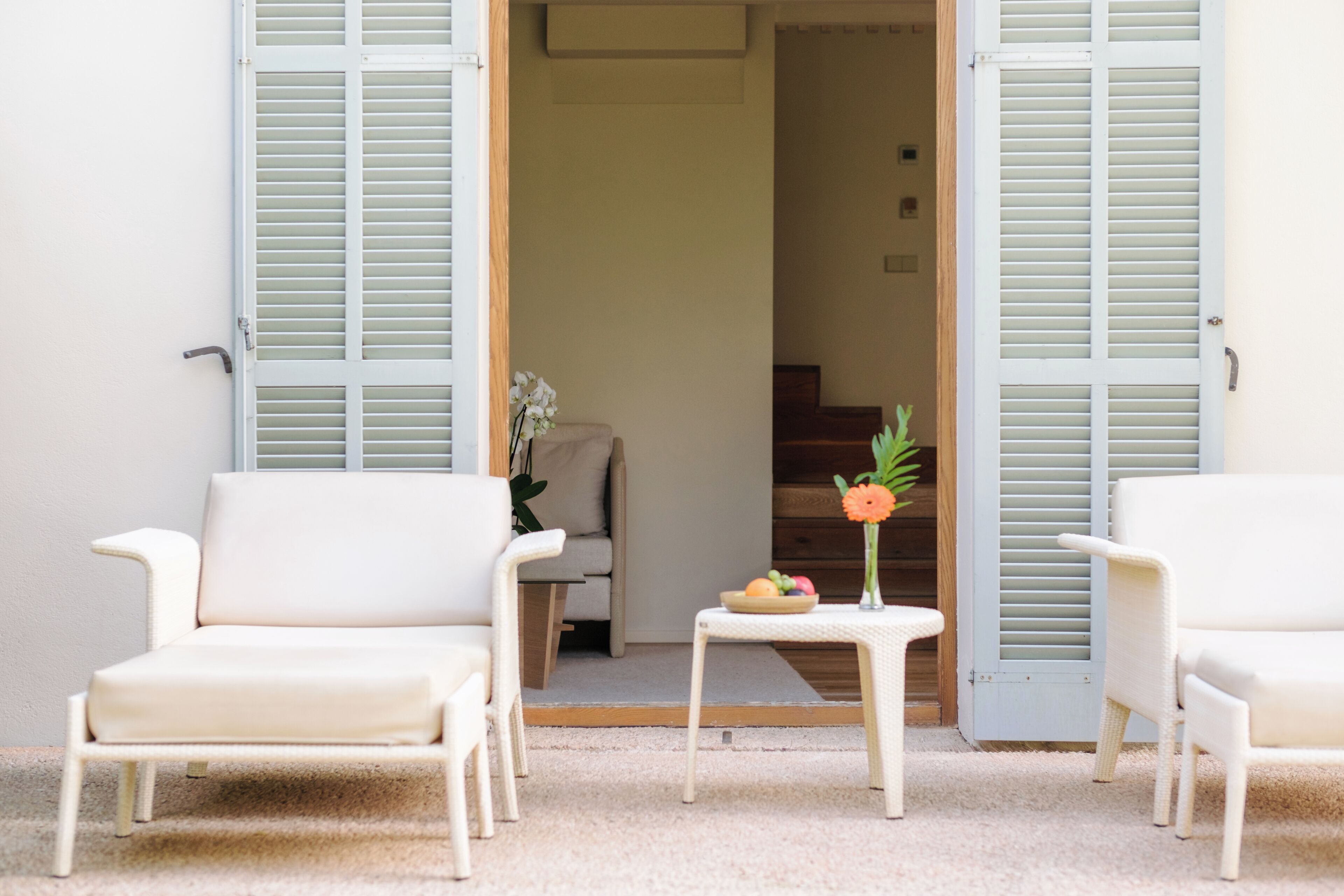 A room with a sofa, two chairs, a small table with a vase and fruit, and a door with shutters.
