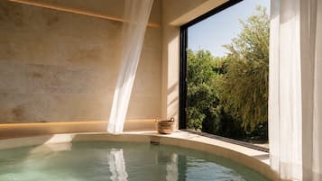 A spa room with a bathtub, a window with curtains, and a view of greenery outside.