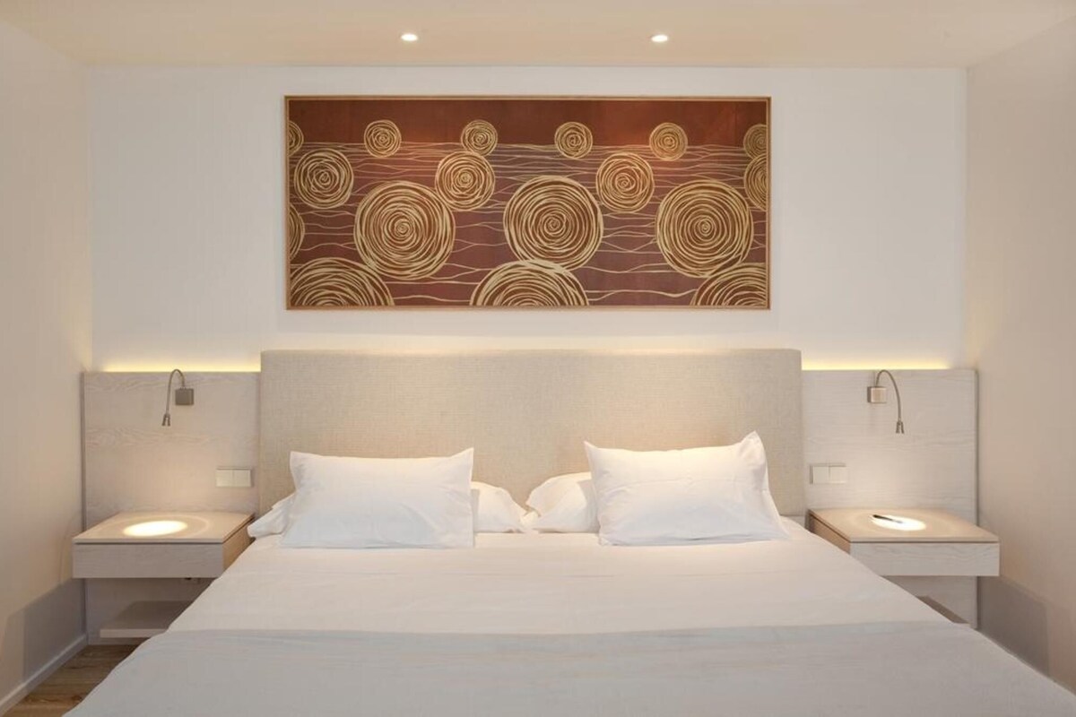 Une chambre d’hôtel avec un grand lit, deux tables de chevet, une œuvre d’art accrochée au mur et un luminaire fixé au plafond.