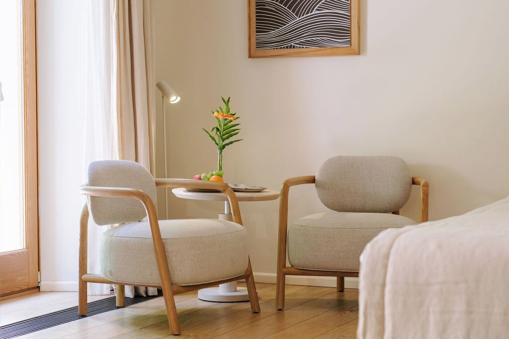 A hotel room with two chairs, a small table, a vase with flowers, and a painting on the wall.