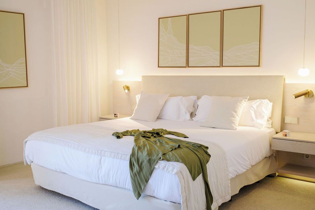 Cama bem arrumada com lençóis brancos e manta verde, luminária de cabeceira e luminária fixada na parede.