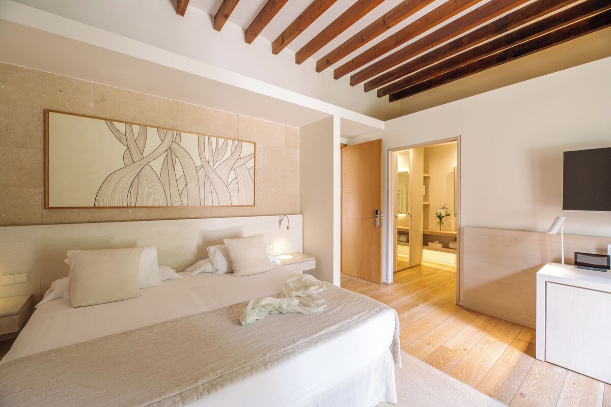 Une chambre moderne avec un grand lit, un plafond en bois, un téléviseur fixé au mur et une porte donnant accès à une salle de bain.