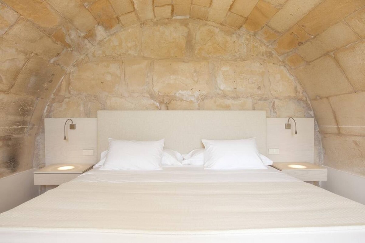 Un lit recouvert de draps blancs se trouve dans une pièce aux murs de pierre, avec deux tables de chevet sur lesquelles reposent des lampes.