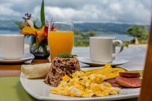 A breakfast plate with rice, eggs, and plantains, accompanied by two cups of orange juice, set against a scenic view of mountains and the sea.