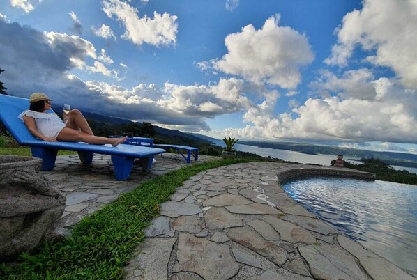 A person relaxing on a blue bench by a stone pathway leading to a pool, with a view of mountains and a lake.