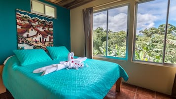 A bed with a turquoise bedspread and pillows, a white towel with a flower, and a painting of a village on the wall.