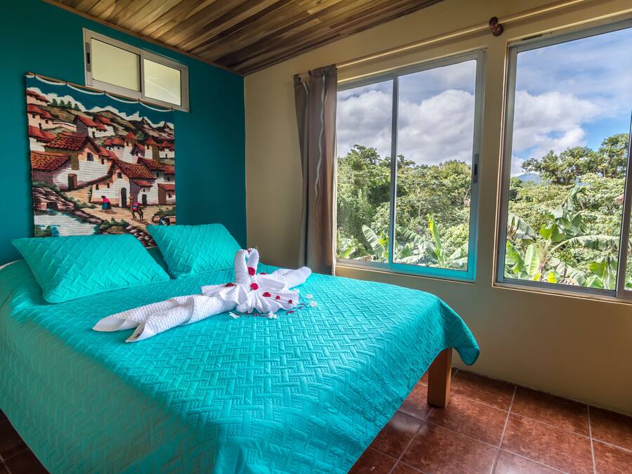 A bed with a turquoise bedspread and pillows, a white towel with a flower, and a painting of a village on the wall.