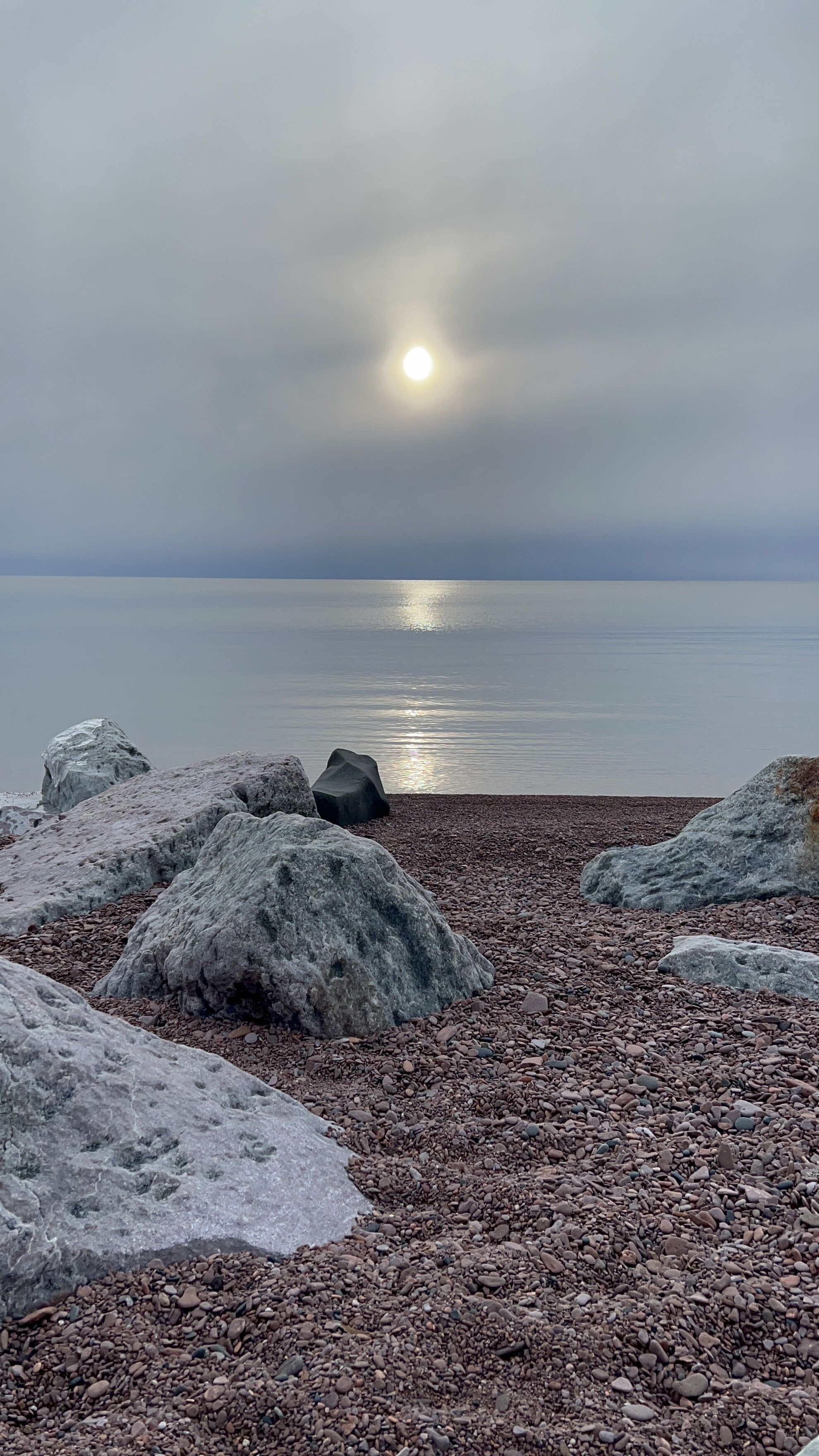 Una spiaggia con ciottoli, grandi rocce e un mare calmo sotto un cielo nuvoloso.