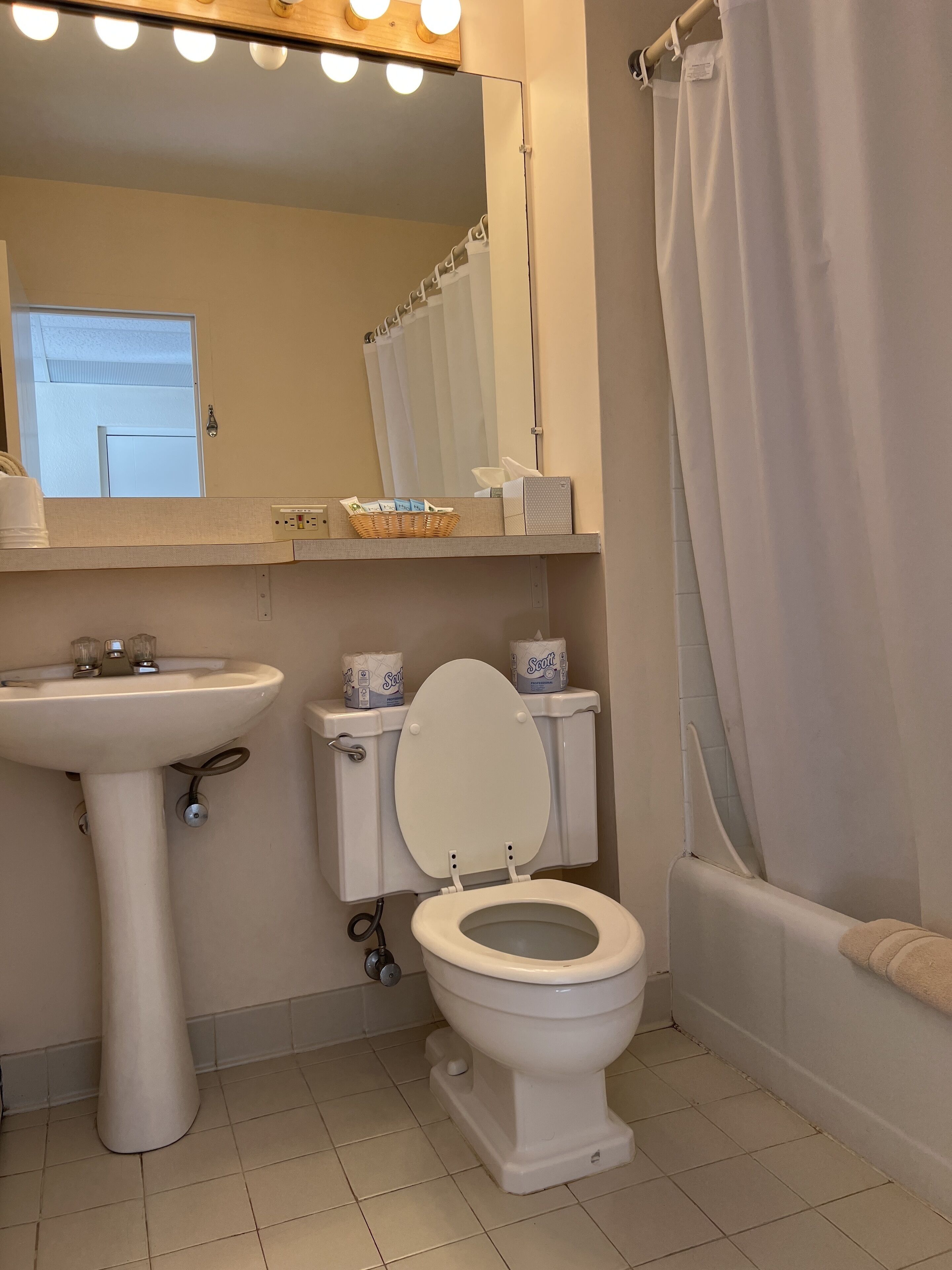 A bathroom with a pedestal sink, toilet, and bathtub. There is a mirror above the sink and a window with blinds.