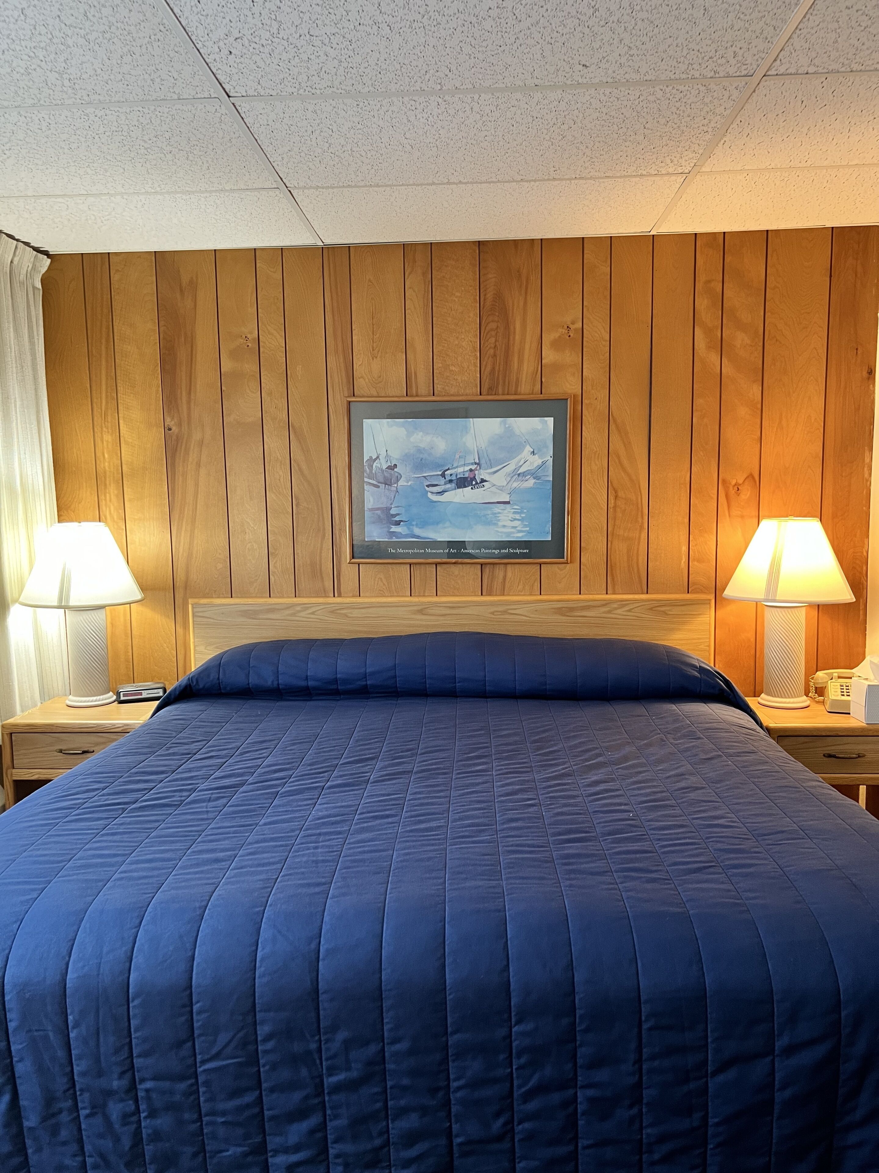 A hotel room with a wooden paneled wall, a framed painting of boats, a large bed with a blue quilt, and two bedside lamps.