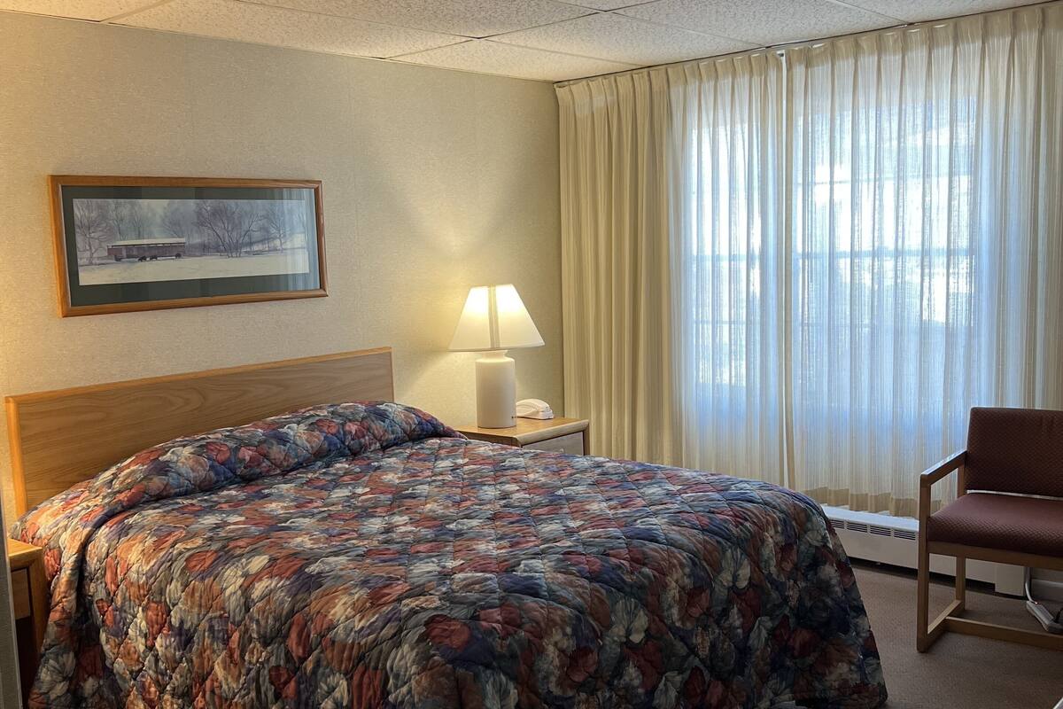 A hotel room with a large bed, a bedside table with a lamp, a chair, a window with curtains, and a framed picture on the wall.