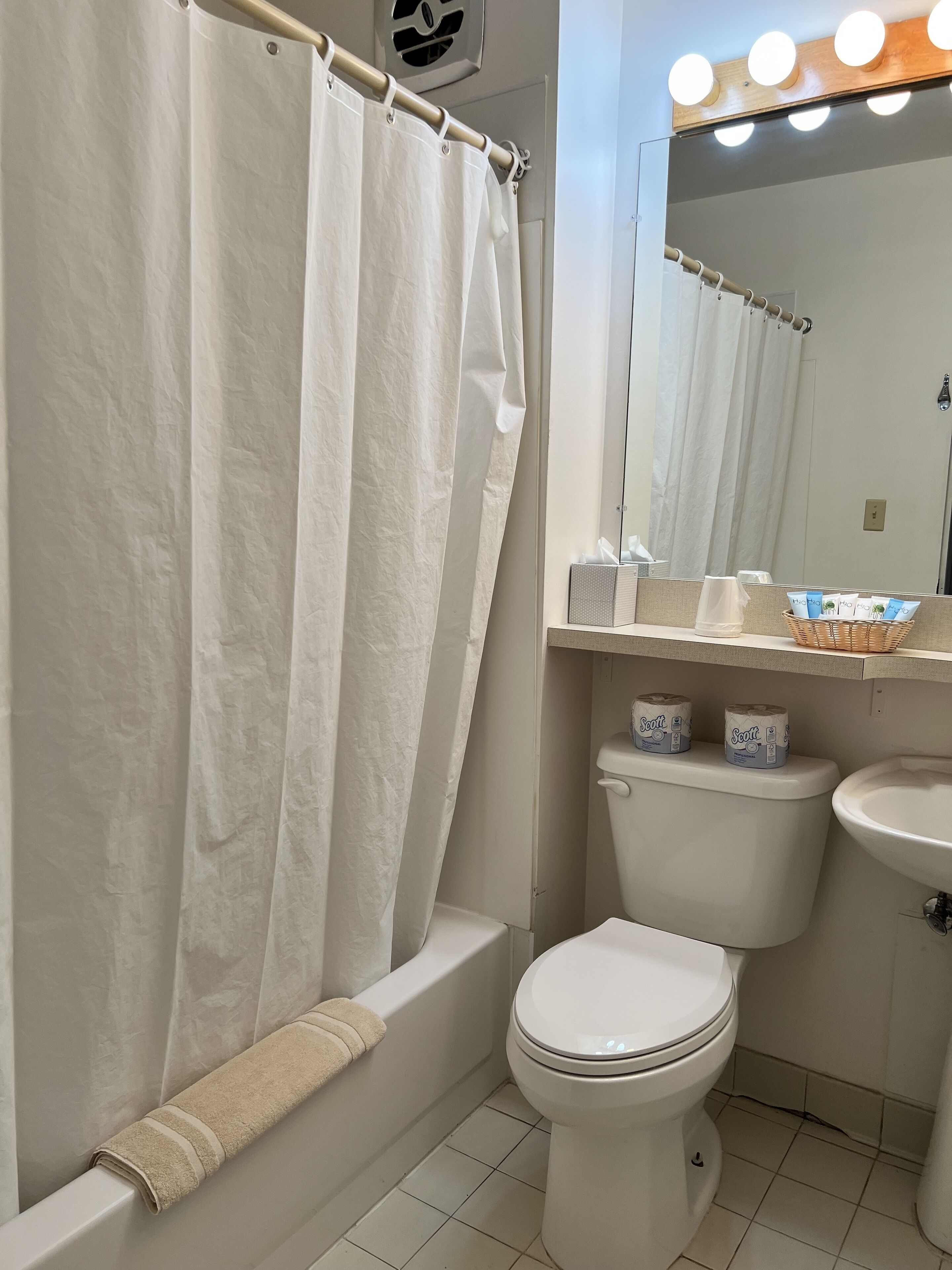 Un baño con inodoro blanco, bañera con toalla color beige, espejo con iluminación y papel higiénico en el tanque.