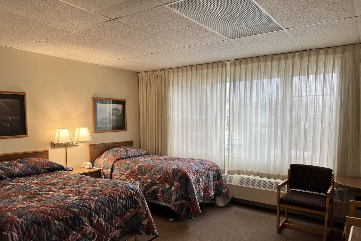 A hotel room with two beds, a chair, a desk, a lamp, and a window with curtains.
