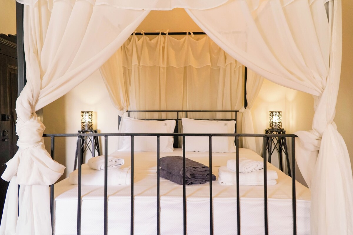 Een hemelbed met witte lakens en twee bedlampjes.