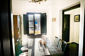 A dining area with a wooden table, chairs, and a view through a window with open shutters. - Santorini Heritage Villas & Mansions (Santorini)